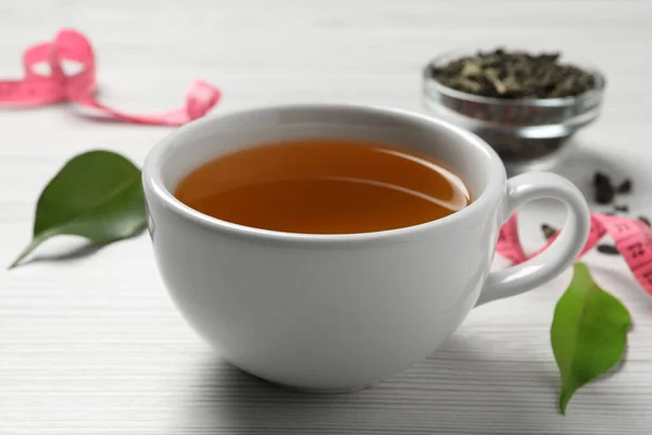 stock image Cup of herbal diet tea, measuring tape and green leaves on white wooden table, closeup. Weight loss concept