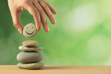 Woman putting stone with drawn happy face onto stack against blurred background, closeup and space for text. Zen concept clipart