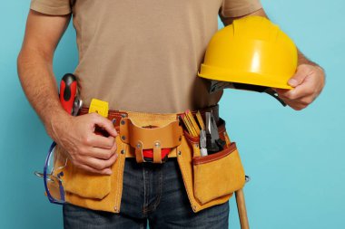 Professional builder with hard hat and tool belt on light blue background, closeup