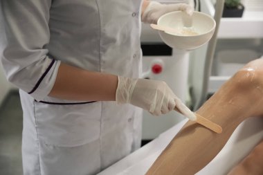 Professional cosmetologist applying gel on client's leg before laser epilation procedure in salon, closeup