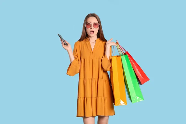 Überraschte Junge Frau Mit Einkaufstaschen Und Smartphone Auf Hellblauem Hintergrund — Stockfoto