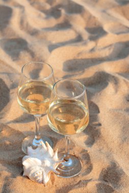 Glasses of tasty wine and seashell on sand, space for text clipart