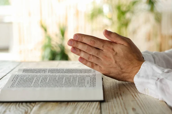 Man with Bible praying at white wooden table indoors, closeup