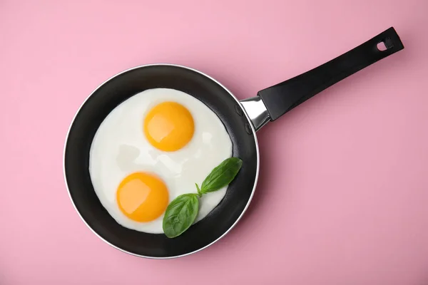 Tasty fried eggs with basil in pan on pink background, top view