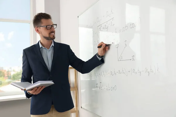 Happy teacher with book explaining mathematics at whiteboard in classroom