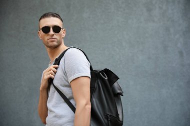 Gri duvarın yanında sırt çantalı, şık güneş gözlüklü yakışıklı genç adam, mesaj için yer var.