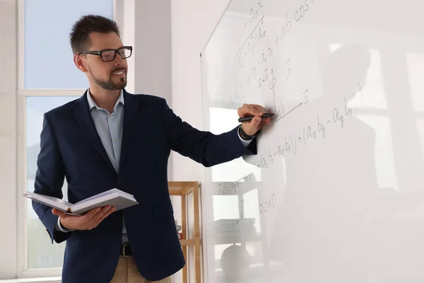 Happy teacher with book explaining mathematics at whiteboard in classroom