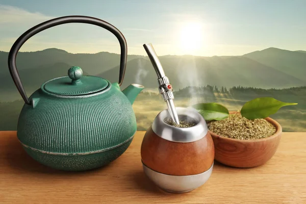 Calabash with mate tea, bombilla and teapot on wooden table and beautiful view of mountain landscape