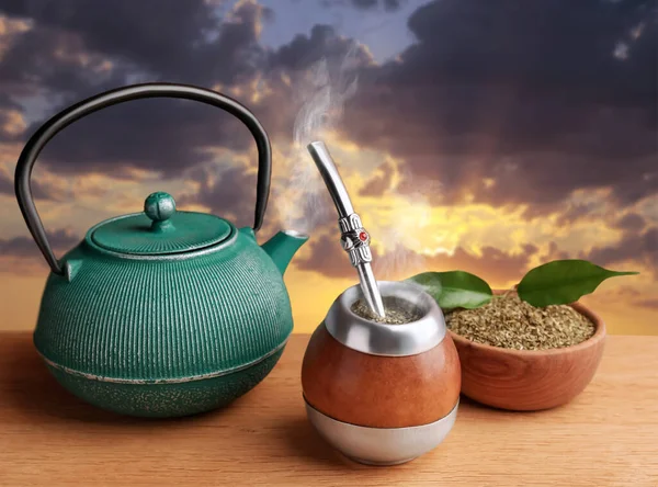 Calabash with mate tea, bombilla and teapot on wooden table outdoors at sunset