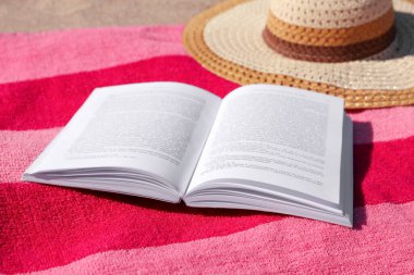 Kumsalda açık kitap, şapka ve çizgili havlu.