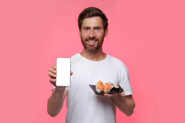 幸せな男はピンクを背景においしい寿司ロールとスマートフォンとプレートを保持 — ストック写真