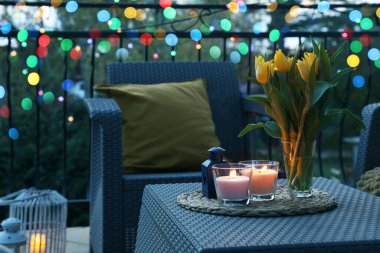 Soft pillows, blanket, burning candles and yellow tulips on rattan garden furniture in evening clipart