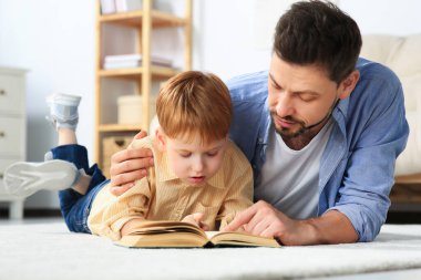 Father reading book with his son on floor in living room at home clipart