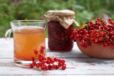 Bir fincan çay, reçel ve olgun viburnum meyveleri açık havada beyaz ahşap masada.