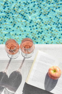 Lezzetli bir gül şarabı, açık bir kitap ve yüzme havuzu kenarında elma.