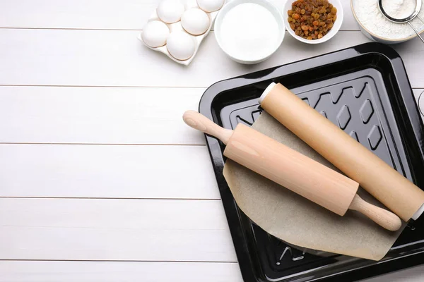 Baking pan with parchment paper, different ingredients and kitchen tools on white wooden table, flat lay. Space for text