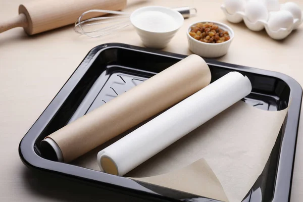 Baking pan with rolls of parchment paper, different ingredients and kitchen tools on wooden table