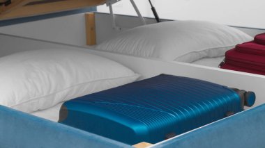 Storage drawer under bed with blue suitcase and white pillows indoors, closeup clipart