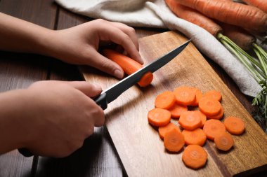 Woman cutting tasty carrot at brown wooden table, closeup