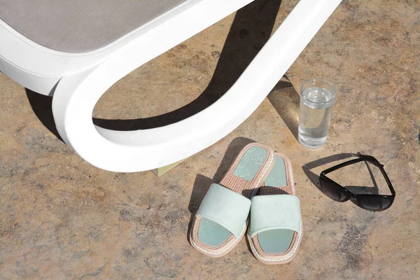 stock image Stylish slippers, sunglasses, glass of water and grey sunbed outdoors. Beach accessories