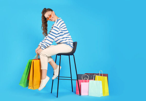 Happy woman in stylish sunglasses holding colorful shopping bags on stool against light blue background