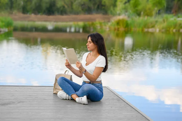 年轻女子在湖边的码头看书 — 图库照片