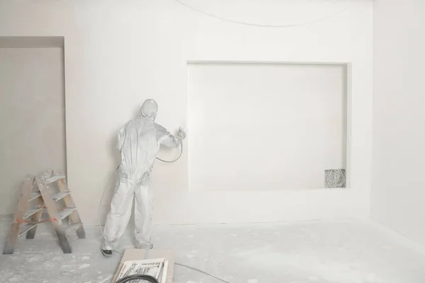 stock image Decorator in protective overalls painting wall with spray gun indoors