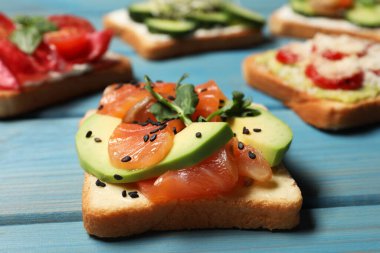 Açık mavi ahşap masada farklı malzemelerle lezzetli tostlar, yakın plan.