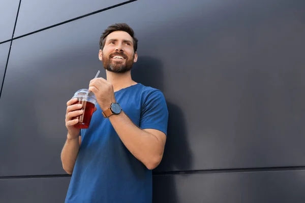 Happy man with delicious juice near grey wall outdoors, low angle view. Space for text