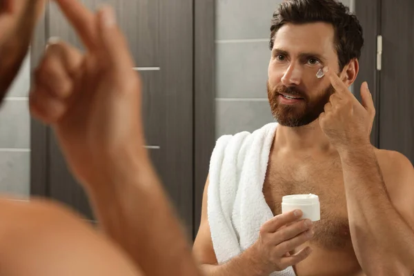 Handsome man applying cream on face in bathroom near mirror