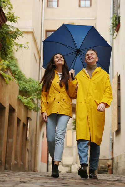 Lovely young couple with umbrella walking under rain on city street, low angle view