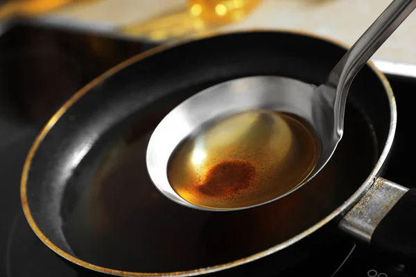 Ladle with used cooking oil over frying pan on stove, closeup