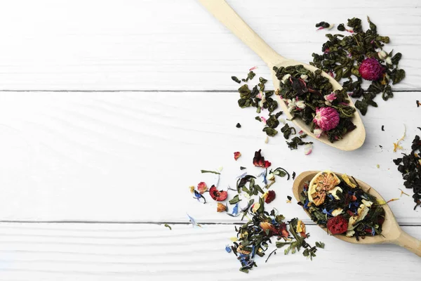 Spoons with dried herbal tea leaves and fruits on white wooden table, flat lay. Space for text