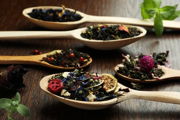 Spoons with dried herbal tea leaves and fruits on wooden table, closeup