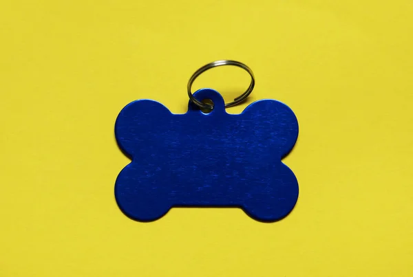 Blue metal pet tag in shape of bone with ring on yellow background, top view. Space for text