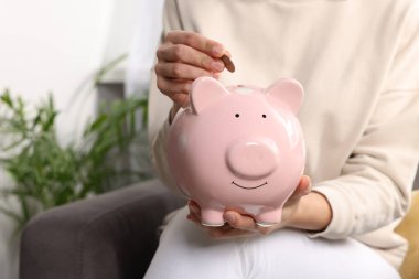 Woman putting coin into piggy bank at home, closeup clipart