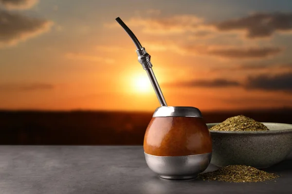 Calabash with mate tea and bombilla on light grey table outdoors at sunset. Space for text
