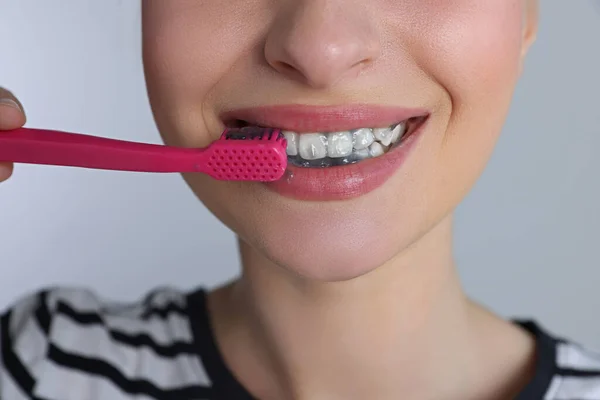 Woman brushing teeth with charcoal toothpaste on grey background, closeup