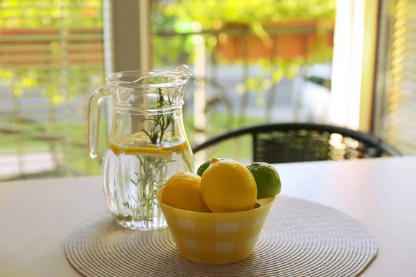 Jug with refreshing lemon water and citrus fruits in bowl on table indoors