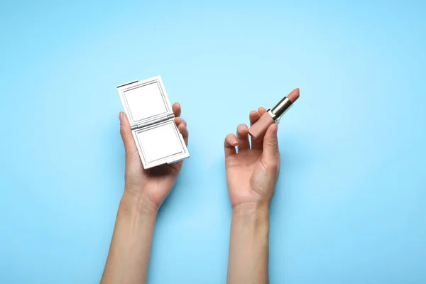 Woman holding pocket mirror and lipstick on light blue background, top view