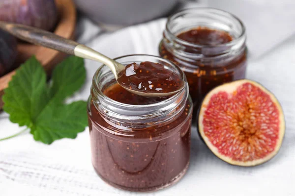 stock image Glass jars of tasty sweet fig jam with spoon and fruits on white wooden table, closeup