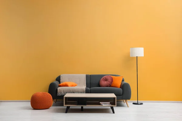 stock image Stylish grey sofa with colorful pillows, wooden table and lamp near pale orange wall indoors. Interior design