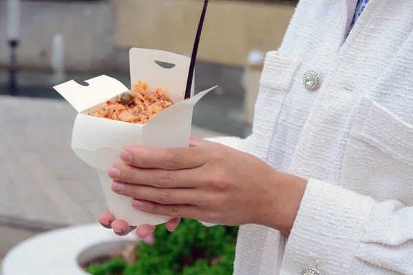 stock image Woman holding paper box of takeaway noodles with fork outdoors, closeup. Street food