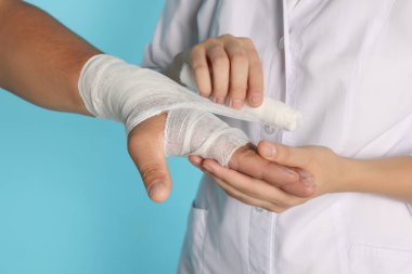 Doktor açık mavi arka planda hastanın eline bandaj uyguluyor.