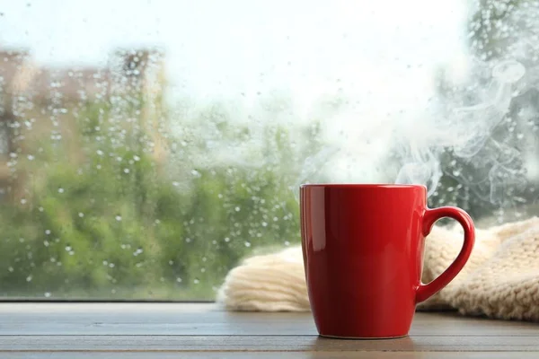 Cup of hot drink and knitted plaid near window on rainy day. Space for text