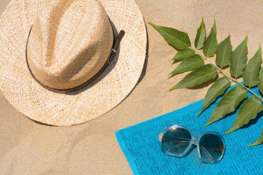 Yumuşak mavi plaj havlusu, güneş gözlüğü, hasır şapka ve kumdaki yeşil yapraklar.