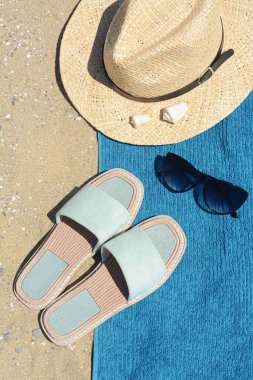 Şık terlikler, hasır şapka, güneş gözlüğü ve kumdaki mavi havlu. Plaj aksesuarları