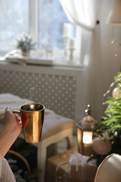 Woman with golden cup of hot drink in room decorated for Christmas, closeup