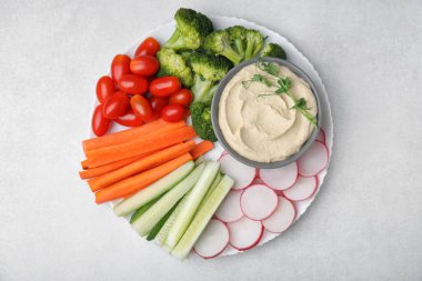 Plate with delicious hummus and fresh vegetables on light grey table, top view clipart