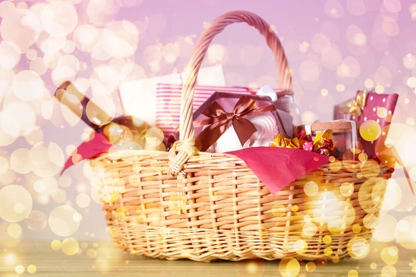 Wicker basket with gifts, wine and food against blurred festive lights. Christmas celebration